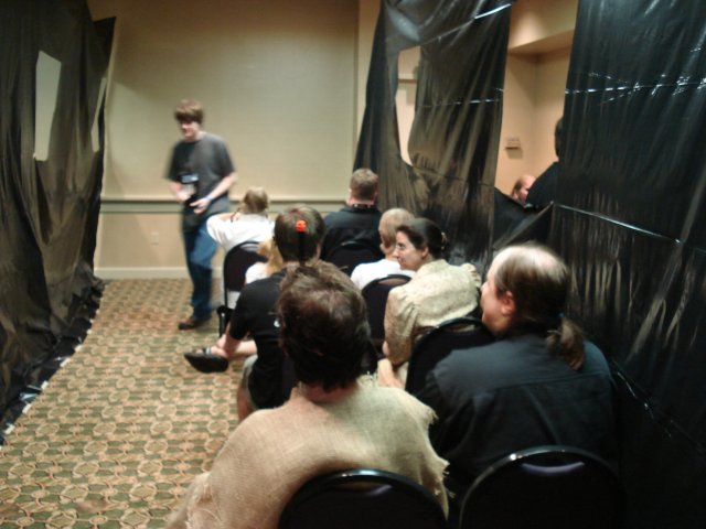 A train car in the Object of Desire game. The walls are marked with black plastic sheets.  Holes cut in the plastic represent windows. Chairs have been arranged into rows and players are sitting in them.
